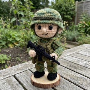 Könnte beinhalten: Gehäkelte Soldat-Puppe mit Tarnhelm und Uniform, die ein schwarzes Gewehr hält. Die Puppe besteht aus beige, grüner und schwarzer Wolle und steht auf einem kleinen Holzsockel. Der Hintergrund ist eine verschwommene Szene im Freien.