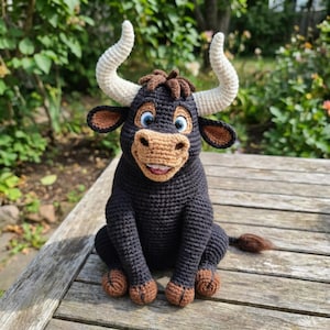 May include: A handmade crochet bull with a friendly expression. The bull features a black body, brown hooves, and a tan muzzle. It has large, curved white horns and blue eyes. The bull is sitting on a wooden surface.