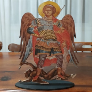 May include: A decorative wooden cutout depicting a figure with wings, a halo, and armor, standing over a fallen figure. The figure is painted with shades of red, gold, blue, and brown. The cutout is mounted on a black base.