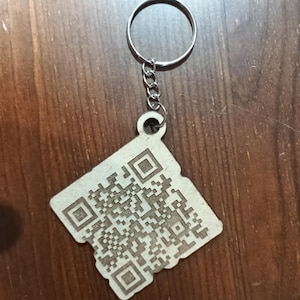 May include: A wooden keychain with a QR code design. The keychain has a silver-colored ring and chain. The QR code is etched into a square-shaped wooden tag.