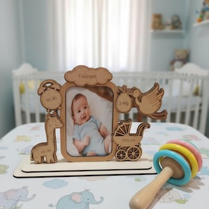 May include: Wooden baby photo frame with a picture of a baby. The frame includes a giraffe, a stork carrying a baby, a baby carriage, and the baby's name. A wooden rattle with colorful rings is in the foreground.