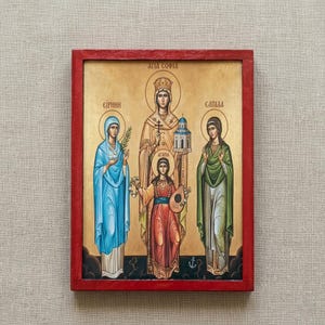 Può includere: Icona religiosa incorniciata con tre figure con aureole su sfondo dorato. Le figure indossano abiti blu, rossi e verdi. La cornice è rossa.
