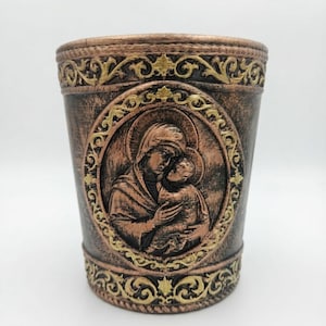 Può includere: Un recipiente cilindrico con finitura bronzo scuro che presenta un rilievo di una figura che tiene in braccio un bambino all'interno di una cornice ovale. I bordi superiore e inferiore sono decorati con motivi in filigrana dorata e la superficie è ruvida.