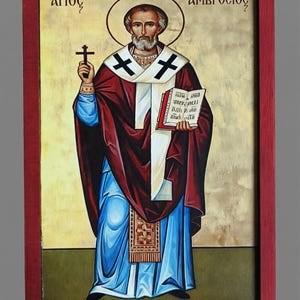 Op de afbeelding: Icoon van een religieuze figuur, omlijst in rood. De figuur draagt een rode mantel, een blauwe jurk en een witte stola met zwarte kruisen. Hij houdt een kruis en een boek vast. De achtergrond is goudkleurig met Griekse tekst.