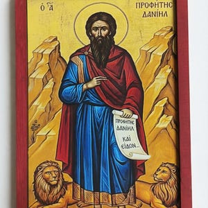 May include: A framed religious icon featuring a central figure with a halo, dressed in blue and red robes, holding a scroll. Two lions are at the bottom. The background is a golden yellow with mountain-like formations. The frame is red.