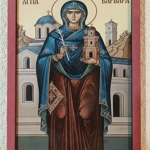 Op de afbeelding: Icoon van Sint Barbara in een rode lijst. De heilige is afgebeeld in een blauwe jurk en bruine jurk, met een toren en een takje. De achtergrond toont een kerk en bergen. De tekst "ΑΓΙΑ ΒΑΡΒΑΡΑ" staat boven de heilige.
