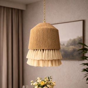 May include: A beige pendant light with a tiered tassel design hangs above a dining table. The light fixture is suspended by a gold chain. A vase of flowers and place settings are on the wooden table.
