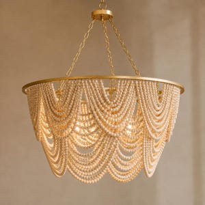 Può includere: Un lampadario dorato con una cornice circolare e fili a cascata di perline di legno bianco sporco. Il lampadario è sospeso a una catena e ha un bagliore caldo e invitante.