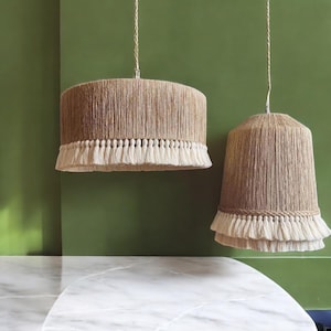 Può includere: Due lampadari sospesi con una trama naturale intrecciata e un bordo di frange bianche. I lampadari sono appesi contro un muro verde.