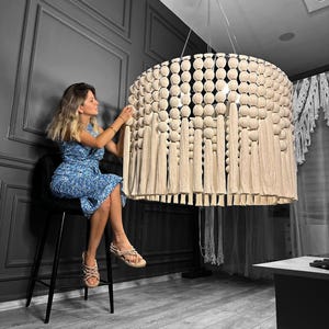 May include: A large, round, cream-colored macrame chandelier with a beaded fringe hanging from the ceiling. A woman is sitting on a chair in front of the chandelier.