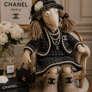 Könnte beinhalten: Eine Stoffpuppe mit Garnhaaren trägt eine schwarz-weiße Tweedjacke, einen passenden Rock und einen Tweedhut mit einer weißen Blume. Die Puppe ist mit Perlenketten, einer Brosche und beigen Stiefeln mit schwarzen Logos geschmückt und hält eine Handtasche.