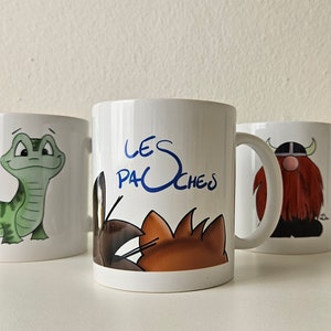 May include: Three white ceramic mugs with cartoon illustrations. The mug on the left features a green lizard. The middle mug has a brown and orange cat and the text "LES PACHEs". The mug on the right has a red-haired viking with a horned helmet.