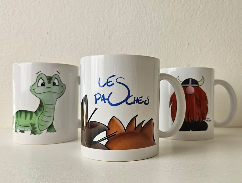 May include: Three white ceramic mugs with cartoon illustrations. The mug on the left features a green lizard. The middle mug has a brown and orange cat and the text "LES PACHEs". The mug on the right has a red-haired viking with a horned helmet.