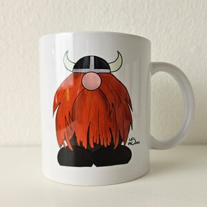 May include: White ceramic mug with a cartoon illustration of a red-haired viking wearing a horned helmet. The mug has a black handle and the artist's signature is visible on the bottom right corner.