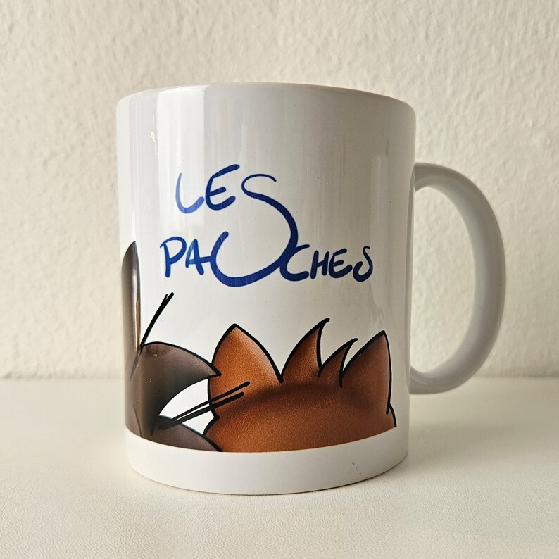 May include: White ceramic mug with a blue and brown cartoon illustration of two cats. The text "LES PACHOCHES" is written in blue on the mug.