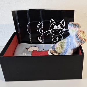 May include: A black box with a red interior, containing a blue and white patterned sock, a white cat and dog illustration, and a white card with black snowflakes.
