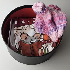May include: A black round gift box with a red interior. The box contains a white tea towel with a cartoon mouse reading a book and a pair of pink and purple knitted socks.