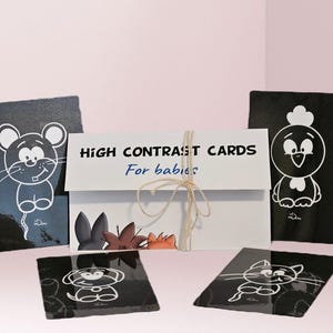 May include: A set of black and white high contrast cards for babies. The cards feature simple illustrations of animals, including a mouse, a chick, a cat, a dog, and a rabbit. The cards are packaged in a white envelope with the text "HIGH CONTRAST CARDS For babies" printed on the front.