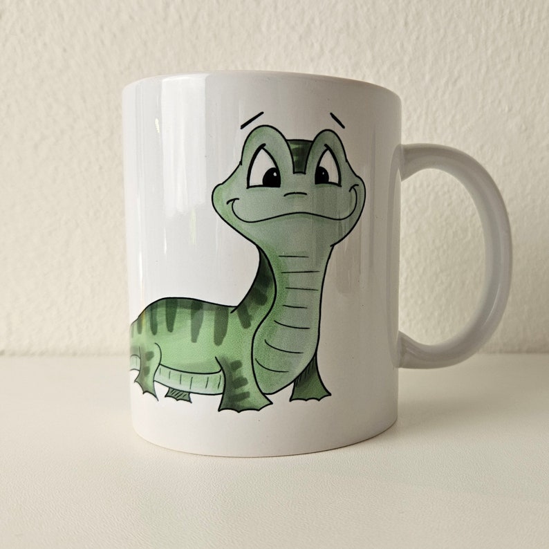 May include: White ceramic mug with a green cartoon lizard with a striped pattern on its back. The lizard is smiling and has large eyes.