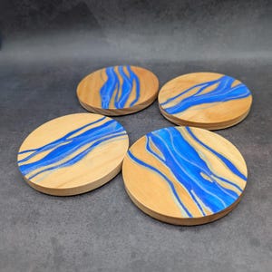 May include: Four round wooden coasters with a natural wood grain and a blue river-like design. The coasters are made of light-colored wood and have a glossy blue resin inlay. The coasters are approximately 4 inches in diameter.