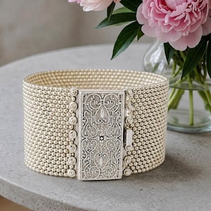 May include: A wide cuff bracelet composed of numerous rows of small, cream-colored beads. The bracelet features a rectangular, silver-toned metal clasp with an intricate, embossed filigree pattern and a central vertical design element.