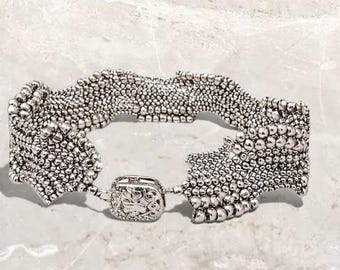 May include: A silver-colored bracelet with a textured, beaded design. The bracelet features a square clasp with an ornate pattern. The overall design is intricate and detailed, with a focus on the texture and shape of the beads.