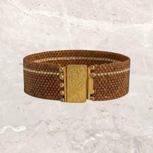 May include: A brown woven bracelet with a gold-colored clasp. The bracelet has a textured appearance with a gold stripe running through the center. The clasp features an ornate design.