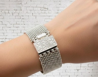 May include: A silver bracelet with a beaded design and an ornate rectangular clasp. The bracelet is composed of small, round beads and features a decorative engraved pattern on the clasp. The bracelet is shown on a wrist.