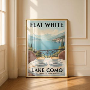 Może przedstawiać: Oprawiony wydruk artystyczny przedstawiający malowniczy widok na jezioro Como we Włoszech, z napisem "FLAT WHITE" u góry i "LAKE COMO" u dołu. Dwie filiżanki kawy stoją na stole z widokiem na jezioro. Wydruk ma złotą ramę.