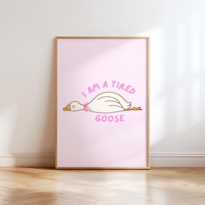 May include: A framed pink art print featuring a cartoon goose lying down, with the text "I AM A TIRED GOOSE" in pink. The goose is white with a yellow beak and a pink bow. The frame is a light brown color.