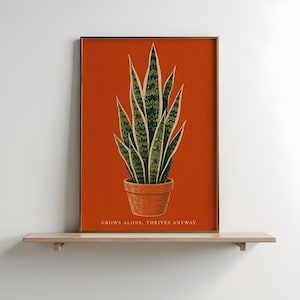 May include: A rectangular wall art print featuring a dark green and light green snake plant in a terracotta pot against a solid burnt orange background. The text "GROWS ALONE. THRIVES ANYWAY." is printed in white at the bottom of the print.