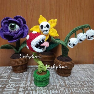 Pode incluir: Figuras de plantas de crochê em vasos. A coleção inclui uma flor roxa com um olho, uma flor amarela com uma caveira, uma planta vermelha e branca com uma boca e uma planta com flores em forma de caveira. O nome do artista, linhpham, é visível.