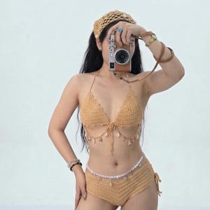 May include: A tan crochet bikini set with a matching headband. The bikini top features seashell accents, and the bottoms have a drawstring tie. A brown camera with a leather strap is held up in front of the face.