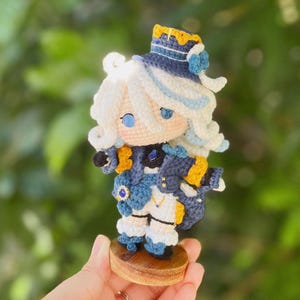 May include: A handcrafted crochet doll with white hair and blue eyes. The doll is dressed in a blue and white outfit with gold detailing and a matching hat. It is mounted on a small wooden base.