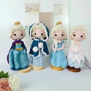 Puede incluir: Cuatro muñecas de ganchillo hechas a mano con cabello de hilo de color claro peinado en trenzas o moños. Cada muñeca lleva un atuendo único en tonos azul, blanco y morado, incluyendo capas, vestidos y capuchas con pompones blancos, sobre bases de madera.