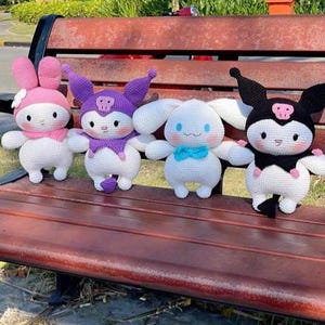 Puede incluir: Cuatro peluches de ganchillo hechos a mano con forma de personajes de dibujos animados están colocados en un banco de madera. Los juguetes son blancos con detalles en rosa, morado, azul y negro. Cada juguete tiene un diseño único, incluyendo sombreros, lazos y rasgos faciales.