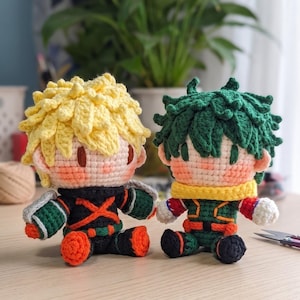 May include: Two handmade crochet dolls featuring anime-inspired designs. The left doll has spiky yellow hair, a black and orange suit with green accents. The right doll has dark green hair, a green and red suit with a yellow scarf around the neck.