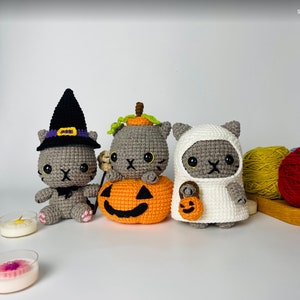 Può includere: Tre giocattoli di gatto in crochet grigi vestiti per Halloween. Un gatto indossa un cappello da strega, un altro è seduto su una zucca arancione in crochet e il terzo indossa un costume da fantasma bianco.