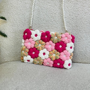 May include: A crocheted floral crossbody bag with a pearl chain strap. The bag is made of pink, white, and green yarn and features a 3D flower design.