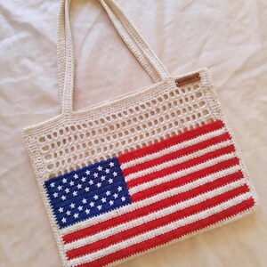 Könnte beinhalten: Eine cremefarbene Häkel-Einkaufstasche mit einem rechteckigen amerikanischen Flaggendesign in Rot, Weiß und Blau auf der unteren Hälfte. Der obere Bereich hat ein offenes Gittermuster und ein kleines braunes Etikett mit der Aufschrift HANDMADE.