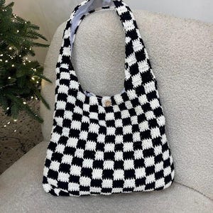 May include: A black and white checkered crochet shoulder bag. The bag has a wide strap and a button closure. The interior lining is white. The bag is sitting on a light gray surface.