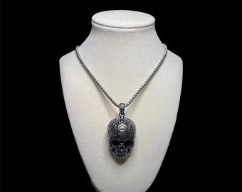 May include: A silver-colored skull pendant necklace. The skull pendant has intricate detailing and is suspended from a matching chain. The necklace is displayed on a white bust against a black background.