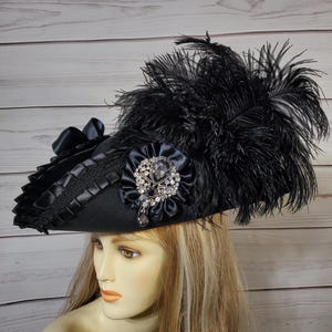 Pu&ograve; includere: Cappello nero con tesa larga, ornato con piume nere, una spilla gioiello e un nastro di raso. Il cappello ha un bordo decorativo attorno alla tesa. Il cappello &egrave; in stile vintage, adatto per costumi o occasioni speciali.