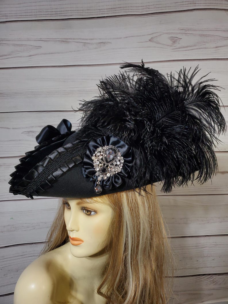 Pu&ograve; includere: Cappello nero con tesa larga, ornato con piume nere, una spilla gioiello e un nastro di raso. Il cappello ha un bordo decorativo attorno alla tesa. Il cappello &egrave; in stile vintage, adatto per costumi o occasioni speciali.