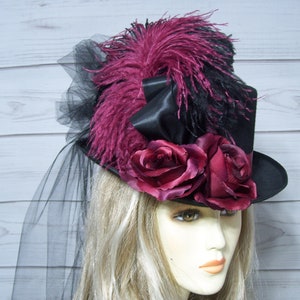 May include: A black top hat with a burgundy feather, black ribbon, and burgundy roses. The hat is adorned with a black veil.