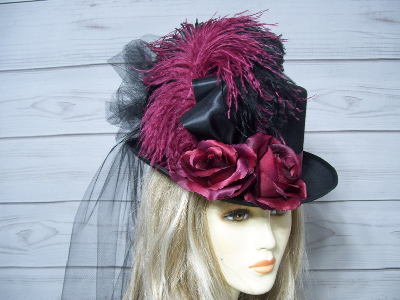 May include: A black top hat with a burgundy feather, black ribbon, and burgundy roses. The hat is adorned with a black veil.