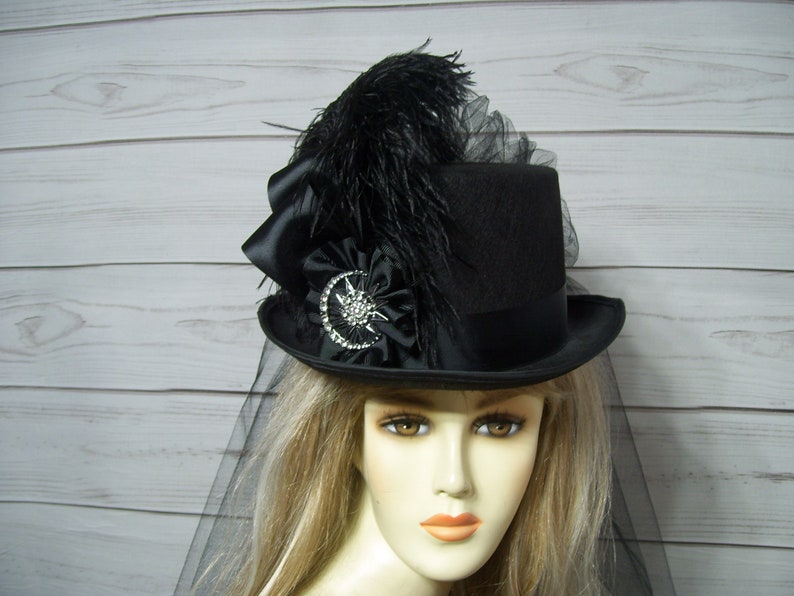 May include: A black top hat with a black feather, a black satin bow, and a jeweled embellishment. The hat is worn by a mannequin with blonde hair and a black veil.