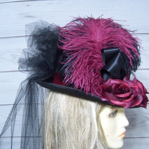 May include: A black top hat with a burgundy velvet band, adorned with a large burgundy rose, black and burgundy feathers, and a black tulle veil.