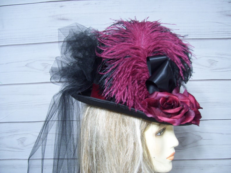 May include: A black top hat with a burgundy velvet band, adorned with a large burgundy rose, black and burgundy feathers, and a black tulle veil.