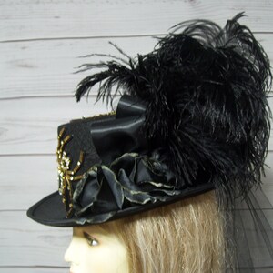 Puede incluir: Un sombrero de copa negro con un dise&ntilde;o de ara&ntilde;a dorado y negro, plumas negras y un velo negro.